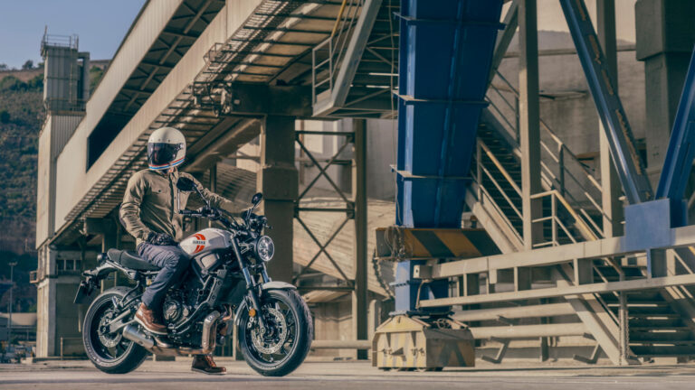 Das Bild zeigt einen Motorradfahrer, der auf einer Yamaha XSR700 sitzt und links aus den Bild hinaussieht. Hinter ihm ist eine Industriebrücke. Ersatzteile und Zubehör für die Yamaha XSR700 gibt es bei Motorradheld.de